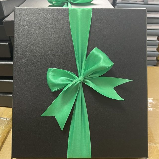 Matte black gift box finished with a green satin ribbon bow, stacked with others behind it.